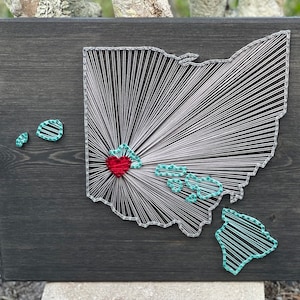 May include: A dark stained wooden board featuring a string art map of Ohio with a red heart. The map is made with white string, with green and white string art islands. The artwork is a unique home decor piece.