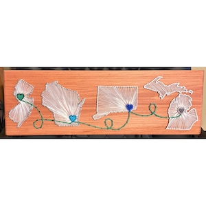 May include: A rectangular wooden board featuring string art of four US state outlines. White string fills the shapes, with small colored hearts placed within each. A continuous green string connects the hearts across the board surface.