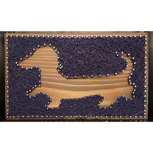 May include: A wooden string art piece featuring a dachshund dog silhouette. The dog shape is cut out of the wood, with navy blue string filling the background. The wooden frame is stained a medium brown color.