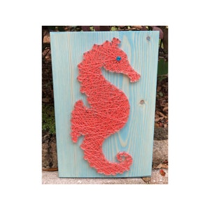 May include: A coral-colored seahorse is created with string art on a light blue wooden background.