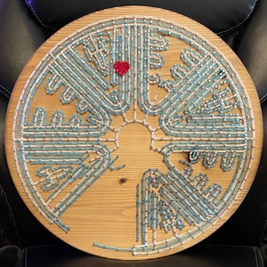 May include: A circular wooden wall art piece with a blue and white geometric design. The design is created with small nails hammered into the wood. A small red heart is in the center of the design.