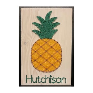 May include: A wooden sign with a string art pineapple design in yellow, green, and brown. The sign has the name "Hutchison" written in green letters at the bottom.