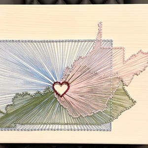 May include: A string art piece on a light wood board featuring the outlines of Kentucky and West Virginia. The states are filled with blue, green, and pink thread radiating from a central red heart shape, all enclosed within a rectangular blue border.
