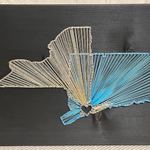 May include: A string art piece depicting the outline of New York State, with a heart shape in the center of the state. The state is made of gold and blue string on a black background.