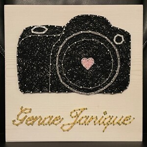 May include: A black and silver string art camera with a pink heart in the lens. The name "Geneva Janique" is written in gold string art below the camera.
