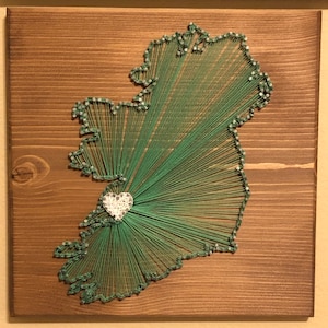 May include: A square wooden board featuring string art in the shape of the island of Ireland. Green thread is wrapped around silver nails outlining the map, with a white heart-shaped cutout placed on the western coast, connected by radiating lines.