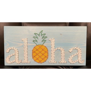 May include: String art of the word "aloha" with a pineapple in the center. The letters are white string on a light blue wooden background.