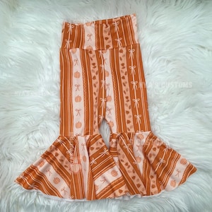 May include: Orange and white striped bell-bottom pants with a fall theme. The pants feature pumpkins, bows, and leaves. The pants have a high waist and a flared leg design. The text "HYA'S CUSTOMS" is visible on the pants.