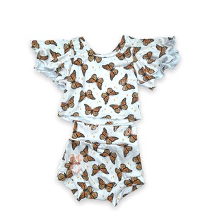 May include: A two-piece baby outfit with a white top and matching shorts. The top has short, ruffled sleeves and a butterfly print. The shorts are also a butterfly print.