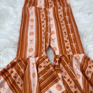 May include: Rust-colored bell-bottom pants with a fall-themed pattern. The pants feature vertical stripes, pumpkins, bows, and leaves in shades of orange and cream. The flared bottoms add a playful touch to the design.
