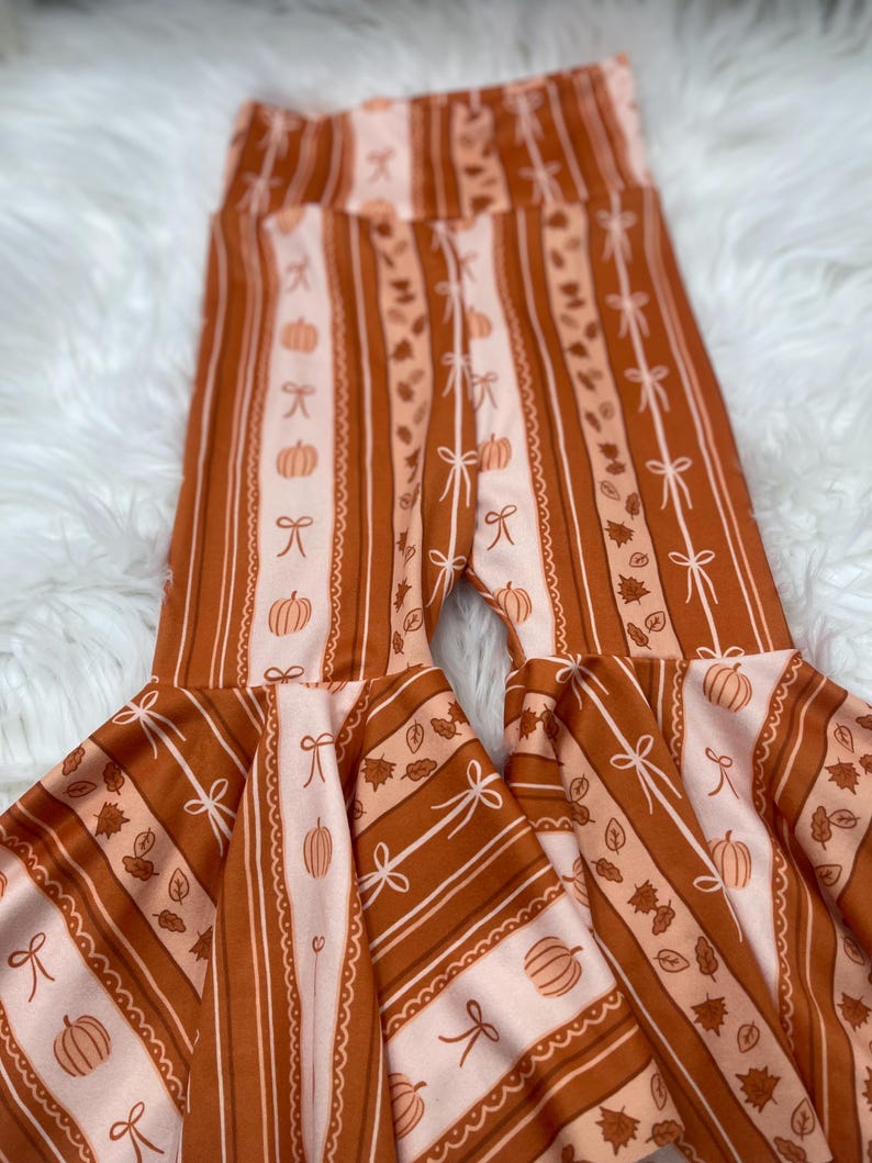 May include: Rust-colored bell-bottom pants with a fall-themed pattern. The pants feature vertical stripes, pumpkins, bows, and leaves in shades of orange and cream. The flared bottoms add a playful touch to the design.