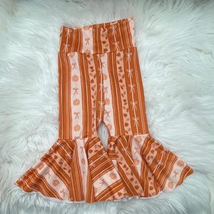 May include: Orange and white striped bell-bottom pants with a fall theme. The pants feature vertical stripes, pumpkins, bows, and leaves. The flared bottoms add a stylish touch to the seasonal design.