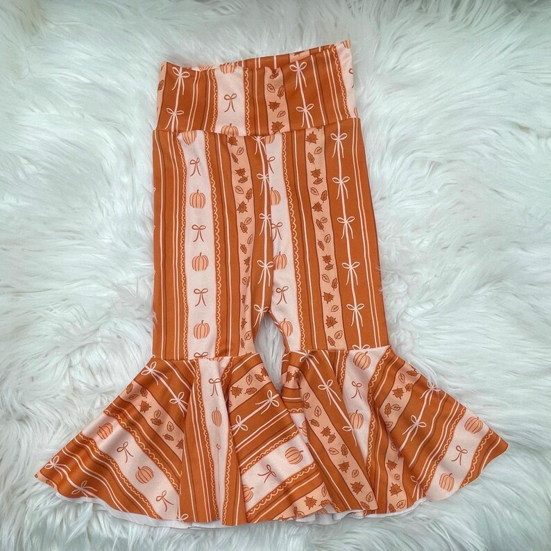 May include: Orange and white striped baby pants with a flared bottom. The pants feature a repeating pattern of pumpkins, bows, and leaves. The fabric has a soft, lightweight appearance, perfect for fall.