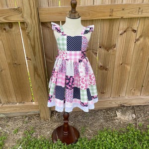 May include: A patchwork dress with a variety of patterns, including floral, polka dot, and gingham, in shades of pink, white, and navy blue. The dress has ruffle sleeves and a white lace trim at the hem. It is displayed on a wooden mannequin.