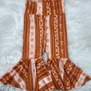 May include: Brown and peach-colored bell-bottom pants with a vertical striped pattern. The stripes alternate between solid brown, peach with white bows, and peach with pumpkins and leaves. The pants have a high waist and flared bottoms.