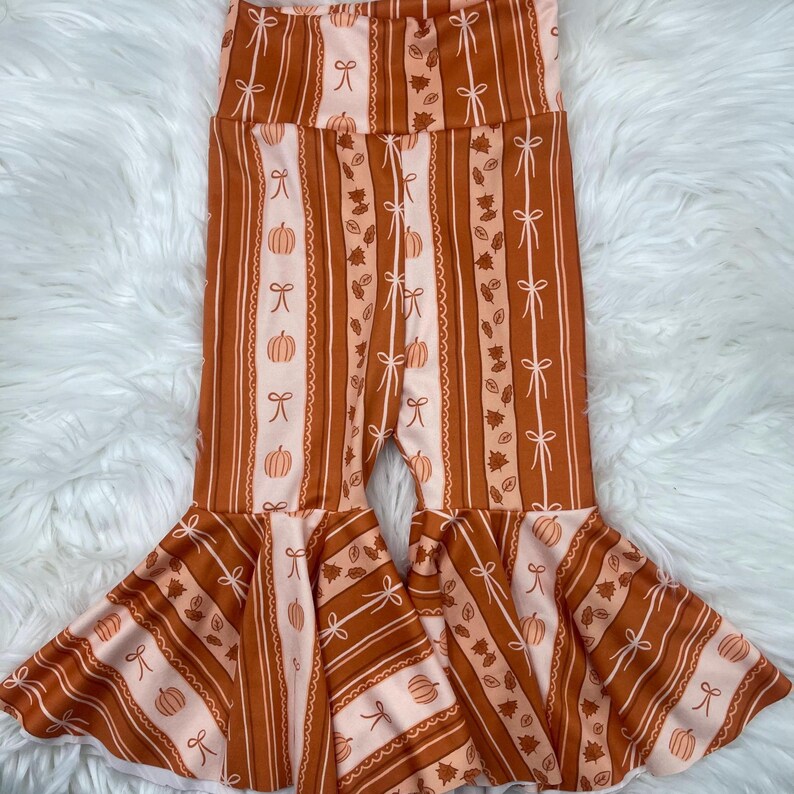 May include: Brown and peach-colored bell-bottom pants with a vertical striped pattern. The stripes alternate between solid brown, peach with white bows, and peach with pumpkins and leaves. The pants have a high waist and flared bottoms.
