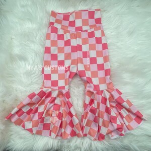 May include: Pink, peach, and white checkered bell-bottom pants. The pants have a high waist and a small pocket on one leg. The text "NYA'S CUSTOMS" is printed on the pants.