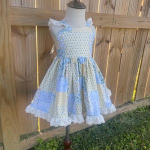 May include: A light blue and yellow patchwork dress with a floral pattern and white lace trim. The dress is displayed on a wooden mannequin and has a square neckline and ruffled sleeves. The skirt is a patchwork of blue and yellow squares.