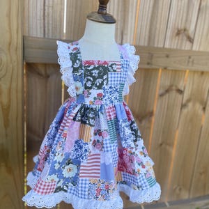 May include: A patchwork-style dress with a square neckline and ruffled sleeves. The dress features a variety of floral, striped, and gingham patterns in shades of pink, blue, green, and white. White lace trim adorns the sleeves and hemline. The dress is displayed on a child-sized mannequin.