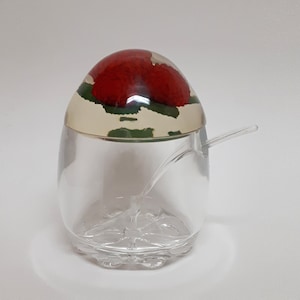 May include: A clear glass sugar dispenser with a red and green floral design on the lid. The dispenser has a small spoon attached to the lid.