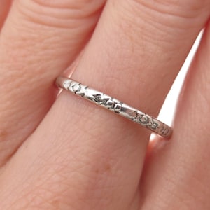 May include: A close-up of a silver ring with a floral pattern. The ring is thin and has a detailed design etched around the band. The ring is worn on a finger.