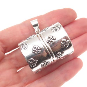 May include: A cylindrical silver pendant featuring a repeating embossed fleur-de-lis pattern across its polished surface. The pendant includes a small silver bail at the top for attachment to a chain, showcasing a metallic finish and detailed relief.