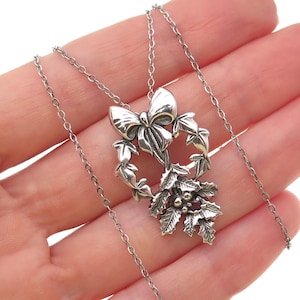 May include: A silver-toned necklace featuring a bow and wreath pendant with holly and berry accents. The pendant is suspended from a delicate chain. The necklace is likely made of sterling silver.