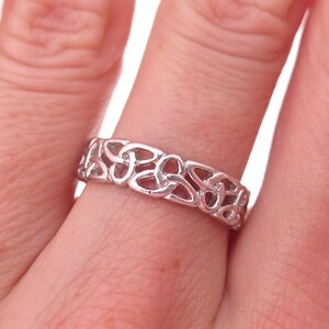 May include: A silver-toned metal ring featuring a continuous band of repeating, interconnected triquetra knotwork patterns. The band has a polished finish and a uniform width, showcasing an intricate, openwork design that wraps around the finger.