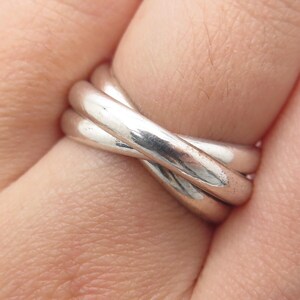 May include: A polished silver ring featuring three interlocking bands that overlap to create a continuous, rounded design. The metal surface is smooth and reflective, showing a bright metallic sheen across the three connected circular loops.