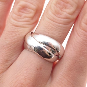 May include: A polished silver-toned ring featuring a triple-band interlocking design. The ring consists of three smooth, rounded metal bands that are woven together to form a single cohesive piece with a reflective, high-shine metallic finish.