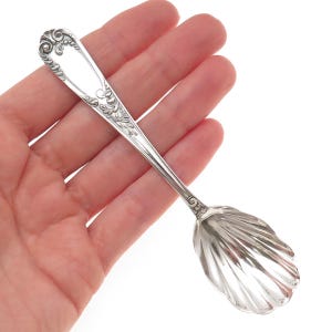 May include: A silver-toned metal spoon featuring a bowl shaped like a scalloped seashell. The handle is decorated with intricate embossed scrollwork and floral-inspired patterns along the edges, tapering toward a smooth, plain central section.