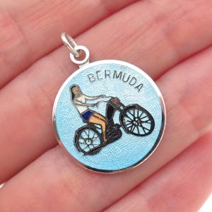 May include: A round silver pendant with a light blue enamel background. The pendant features the word "BERMUDA" in black letters and a person riding a black scooter. The person is wearing white and blue clothing.