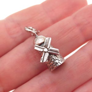 May include: A silver-toned metal charm shaped like a windmill, featuring a rounded top, a central rotating-style blade assembly, and a textured, scale-like pattern on the conical base, complete with a small jump ring attached at the very top.