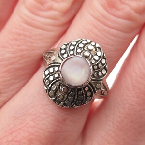 May include: A silver ring with a central, round, pale pink gemstone. The ring's setting features a detailed, swirling design with small, circular accents. The ring is on a finger.