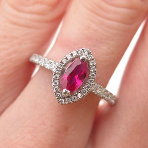 May include: A silver-toned ring featuring a central marquise-cut vibrant pink gemstone surrounded by a halo of small clear stones. The band is also encrusted with small clear stones along the sides, creating a sparkling and detailed appearance.
