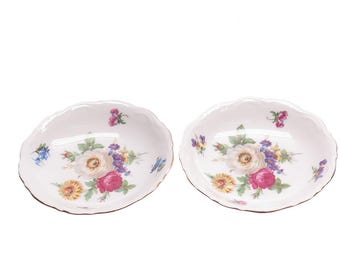 May include: Two white porcelain bowls with scalloped edges and a floral design. The bowls feature a bouquet of pink, white, and yellow flowers with green leaves.