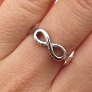 May include: A silver-colored ring featuring an infinity symbol design. The ring is a simple band with the infinity symbol as the focal point, showcasing a minimalist style. The ring is worn on a finger.