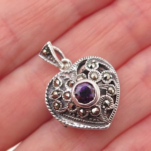 May include: A heart-shaped silver pendant featuring a central round purple gemstone surrounded by intricate filigree metalwork and small embedded marcasite stones. The pendant includes a triangular bail at the top for attachment to a chain.