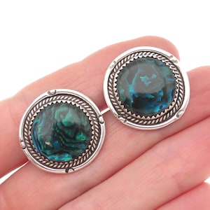 May include: A pair of round silver cufflinks with iridescent abalone shell centers. The abalone shell displays shades of green and blue. The cufflinks have a decorative rope-like border and a scalloped edge around the shell.