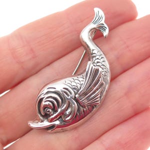 May include: A silver-toned fish brooch with detailed scales, fins, and a curved tail. The brooch has a pin on the back for secure fastening. The fish is depicted in a stylized, artistic manner, making it a unique accessory.