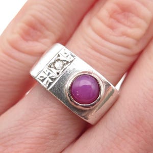 May include: A silver ring with a rectangular design. The ring features a round, deep pink gemstone and a small, clear diamond. The band has a geometric pattern. A piece of jewelry.