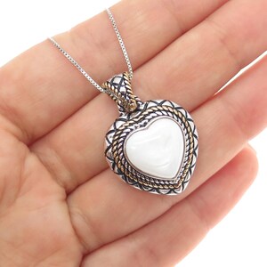 May include: A silver necklace with a heart-shaped pendant. The pendant has a white center, a silver border, and gold accents. The necklace is held in a hand.