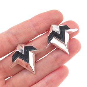 May include: A pair of silver-toned earrings featuring a sharp, angular, geometric star-like shape with recessed black detailing. The earrings have a polished metallic finish and a pointed, multi-faceted design that creates a distinct, bold appearance.