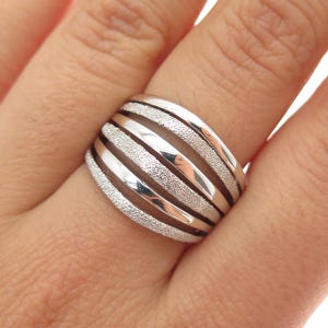 May include: A wide silver ring with a textured, frosted band design. The ring features alternating bands of smooth, polished silver and textured, glittery silver. The ring is worn on a finger.