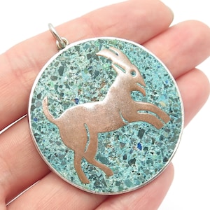 May include: A round pendant with a copper-colored goat silhouette on a turquoise mosaic background. The pendant has a silver-colored border and a small loop at the top for a chain. The goat is depicted in a leaping pose.