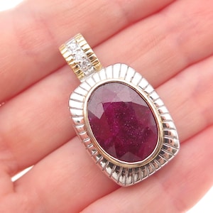 May include: A rectangular pendant featuring a large, oval, faceted deep red ruby set in a gold bezel. The surrounding frame is made of textured silver with vertical grooves. The bail is accented with small, clear, sparkling white stones.
