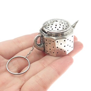 May include: A silver-toned tea infuser shaped like a small teapot. It features a lid with perforations, a handle, and a spout. A chain connects the infuser to a ring. The infuser is held in a hand against a white background.