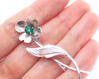May include: A silver-toned floral brooch featuring a flower with four petals and a central green gemstone. The brooch has a stem and a leaf design, with a pin clasp on the back. The brooch is held in a hand.