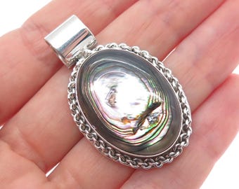 May include: Oval pendant with a polished abalone shell center, framed by a silver chain-link border. The iridescent shell displays shades of green, blue, and purple. The pendant has a silver bail for a necklace.