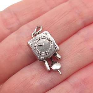 May include: A small, silver-toned clock charm with a square face and a circular clock face. The clock has two small, rectangular weights and a pendulum. The charm is held by a hand.